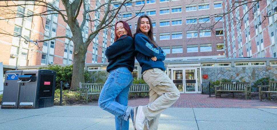 Photo: Sarah Rho (left) and Sarah Kirk poses back to back with arms crossed. Each holds their back foot against the other as the look towards the camera. The both stand in front of a large, brick, tower-style dorm residence.