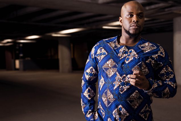 Photo: Patrick Dailey, a bald black man wearing an ornately designed sweater, poses for a portrait in a parking garage.
