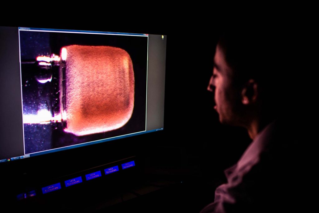 Photo of Christos Michas sitting in front of a large screen in a darkened room. On screen, a magnified image of the heart pump, which is shaped like a gum drop and is pinkish colored. Below, glowing blue rectangular buttons are seen.