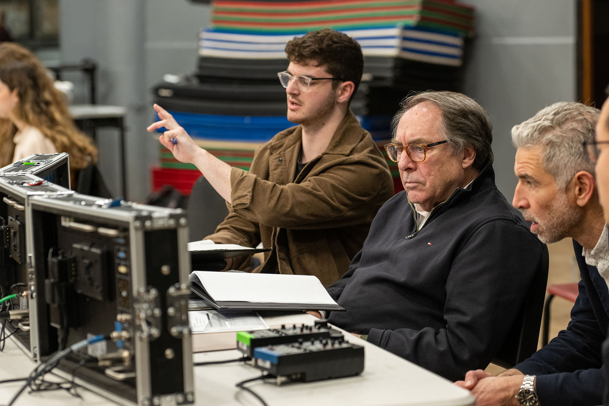 Photo: From left, director Eli Canter (COM’23), Paul Schneider, COM professor of film and television and department chair,  and Tim Palmer, a COM professor of the practice in cinematography, discuss camera blocking during a rehearsal. A young man wearing glasses and a brown collared shirt points forward and speaks as an older man wearing glasses and a black collared shirt sits to his right. They both sit at a table with multiple mini screens in front of them.