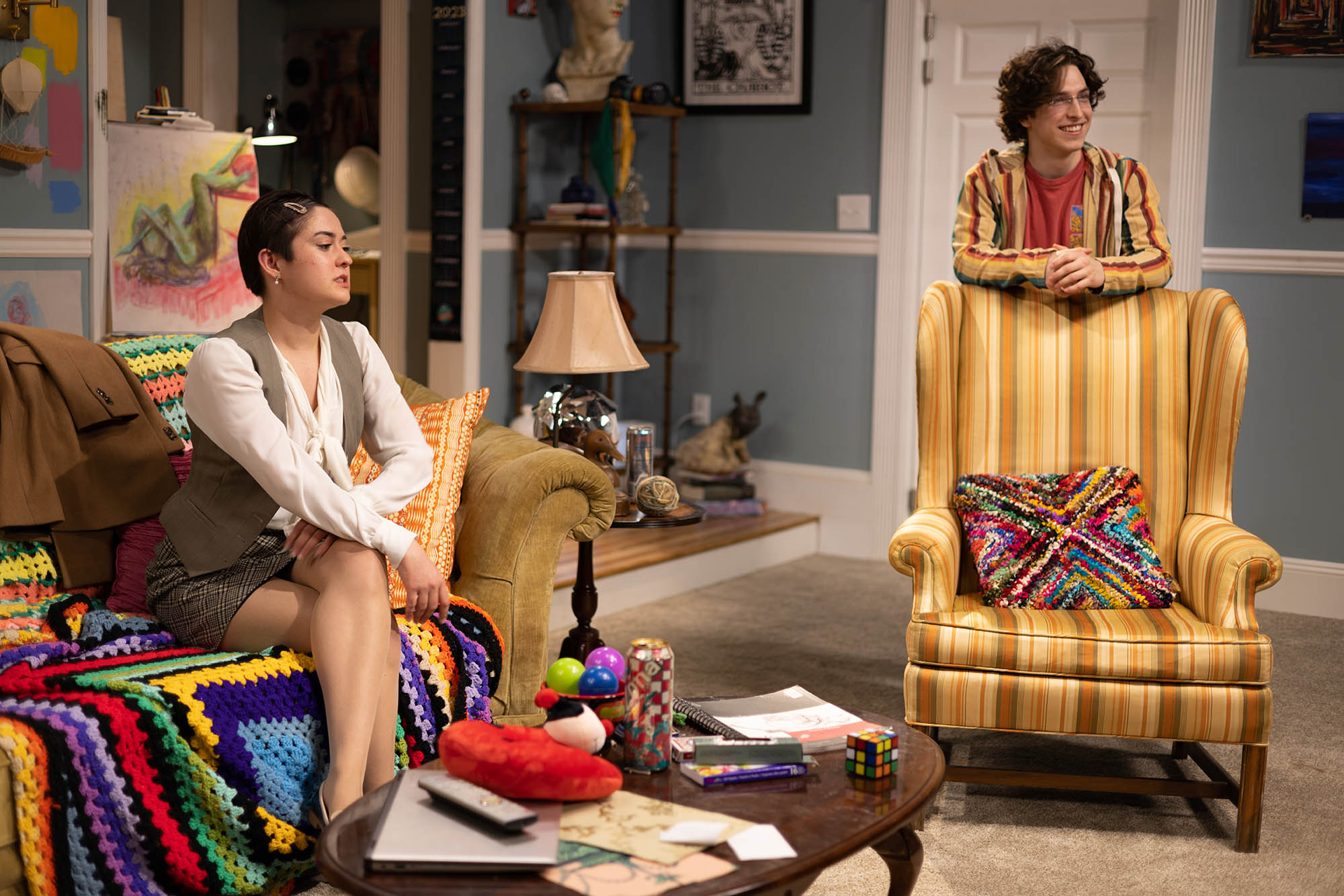Photo: Phil (Right) and Barb Shipley, played by Tyler Statkevicus (CFA’23) and Acsa Welker (CFA’23) look to the right as they chat and smile. Phil stands and leans on the back of a large yellow armchair as Barb sits on an olive green couch and sneers.