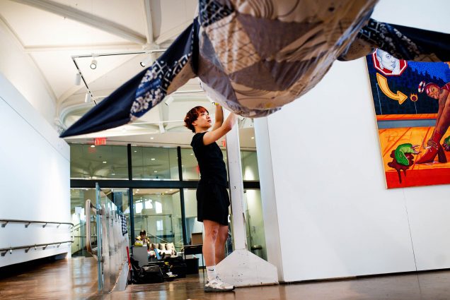 Photo: A young man wearing a black shirt and shorts works on a large sculpture being displayed in an art gallery.