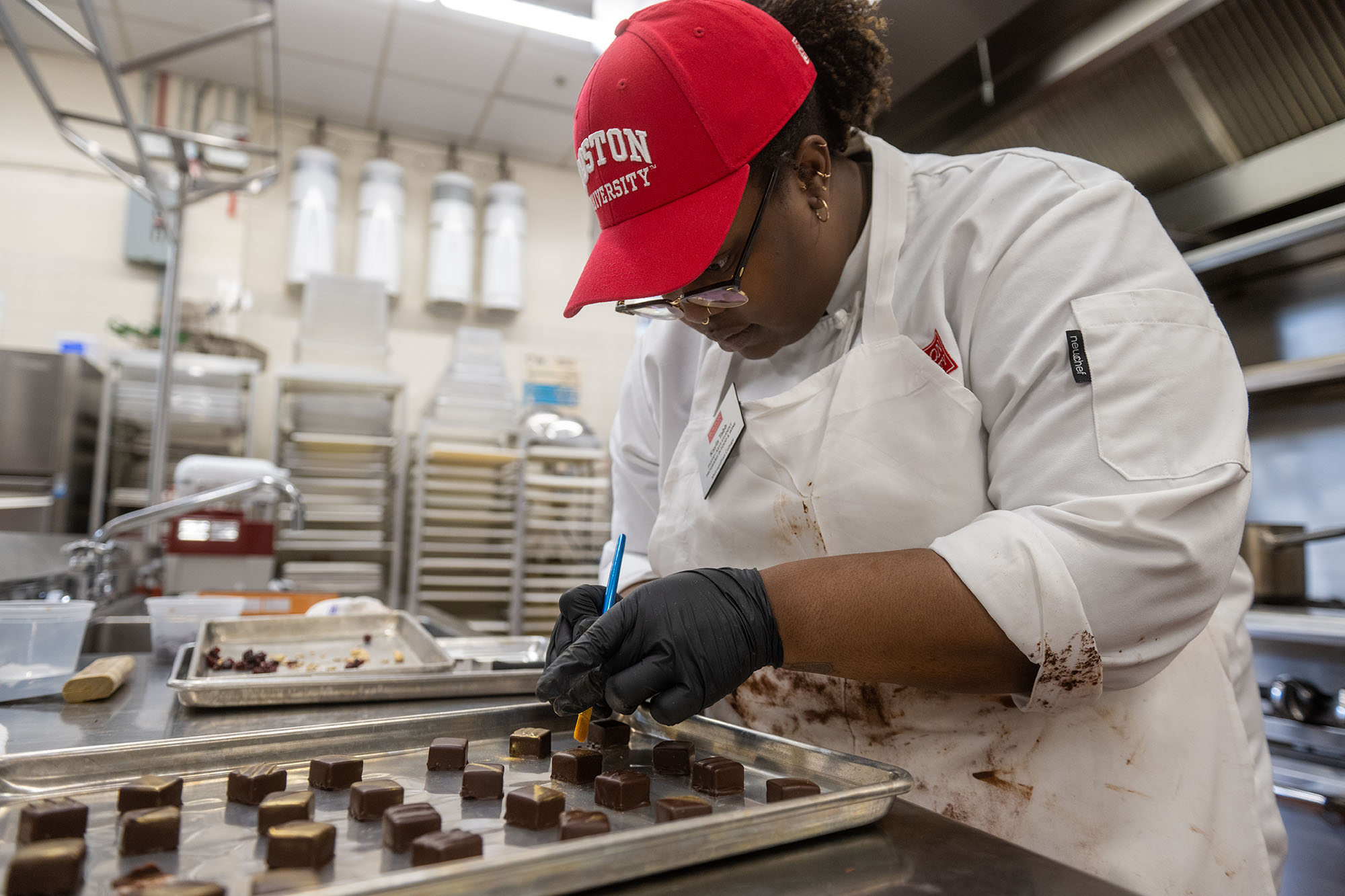 Photo: Kayla Tabb, a young Black woman with hair tied back in a low puff and wearing glasses, a red BU cap, and white chef's outfit, decorates her chocolate covered caramels during the chocolate work class.