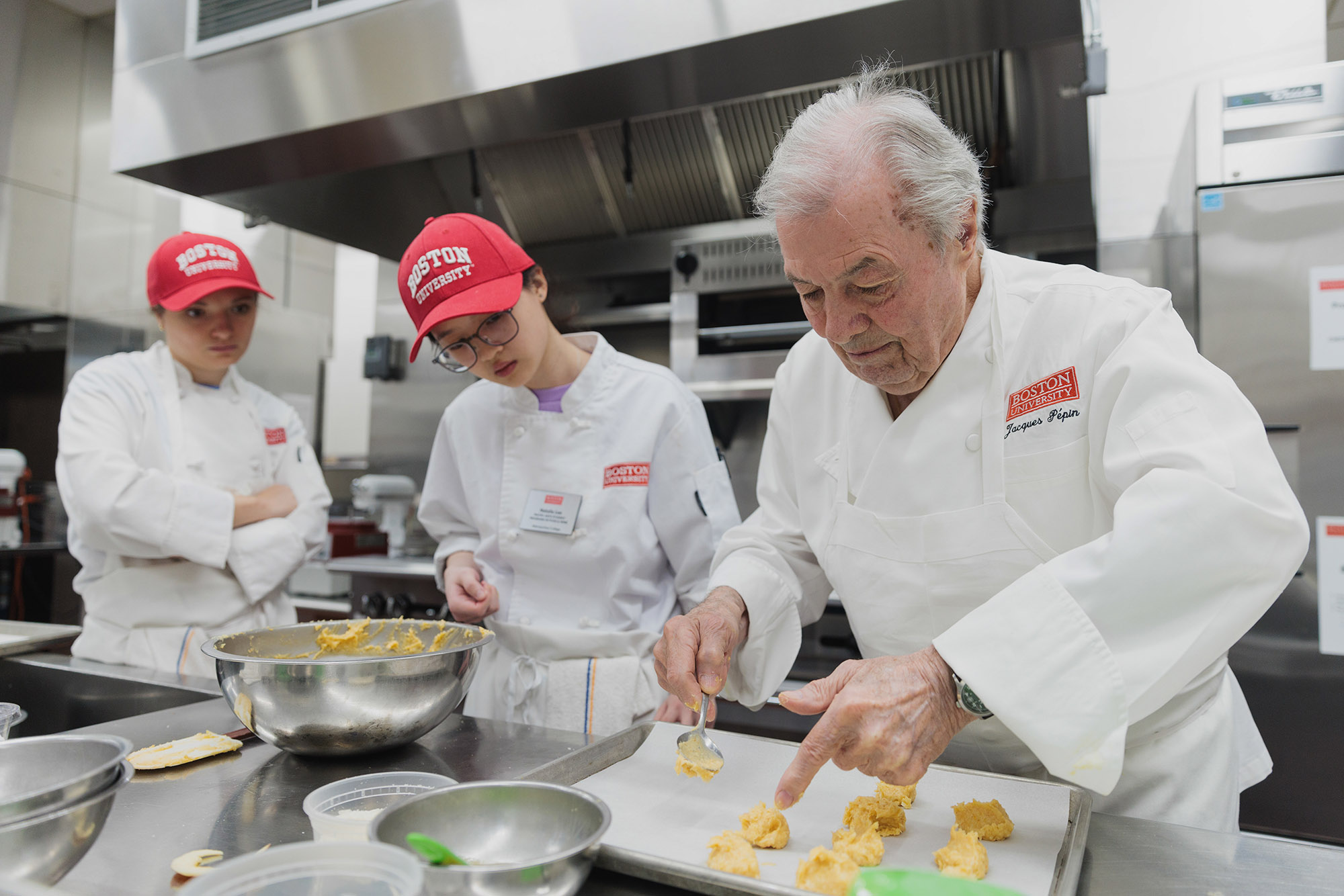 Photo: Mia Ball (MET'23) and Natalie Lee (MET'24) watch Jacques Pepin teach during the Masterclass at the BU Gastronomy Kitchens. Two young women wearing red BU caps and white chef's outfits watch an older white man with silver white hair and wearing a white chef's jacket with his name sewn onto the breast pocket, as he places down choux pastry dough.