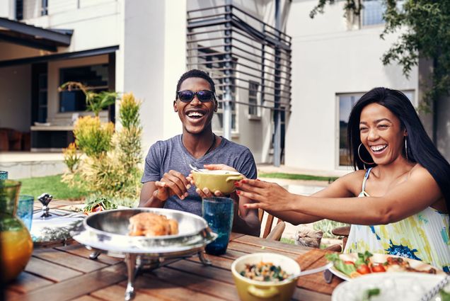 Photo: A stock image of a family enjoying a lunch outside on a wooden table. In the middle, a Black man smiling wide with black shades and a gray t-shirt. He reaches for the food in front of him. Next to him, a Black woman with long hair smiles to something off-camera. She reaches over the table with a bowl in her right hand. The day is bright and sunny.