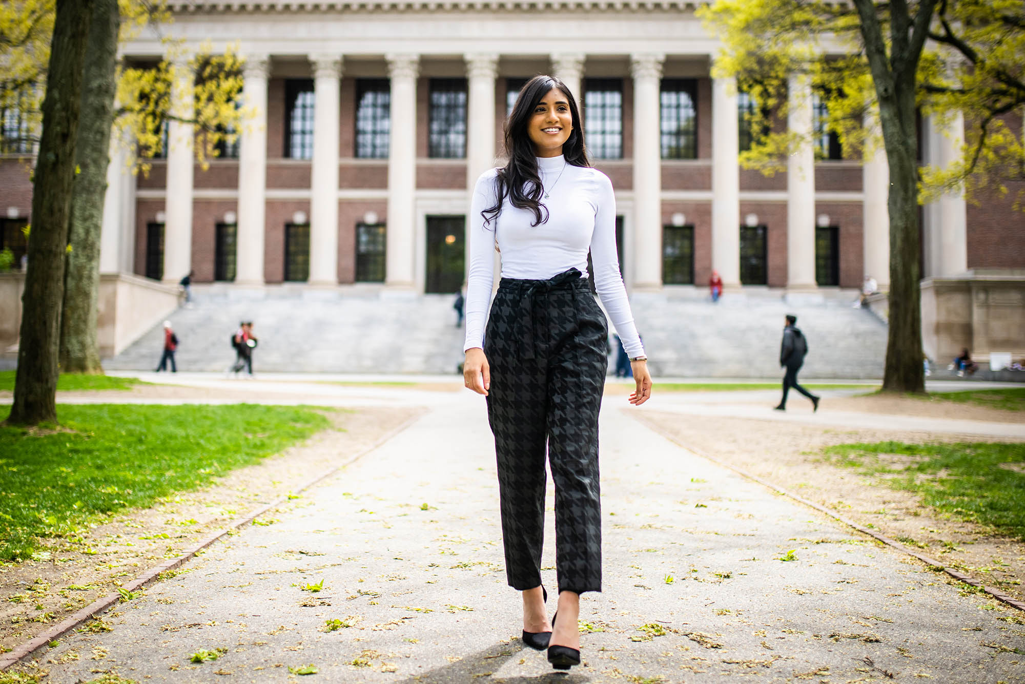 Photo: A Western Asian woman with long, dark hair struts down a path at Harvard. She wears a white turtleneck, plaid pants, and smiles towards something off camera.