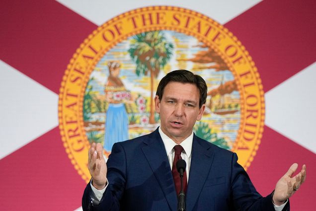 Photo: Ron DeSantis, a white man, stands in front of a banner with Florida's emblem addressing a crowd. He wears a basic navy suit with a red tie.