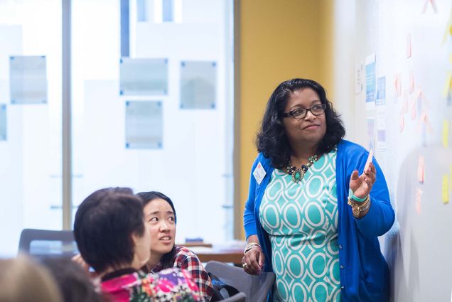 Photo of Shiney James, a brown-skinned woman with short black hair, pointing at a white board. She wears a blue sweater and dress with a colorful white and teal pattern. She wears a name tag and stands the front of the classroom during a section of FY101.