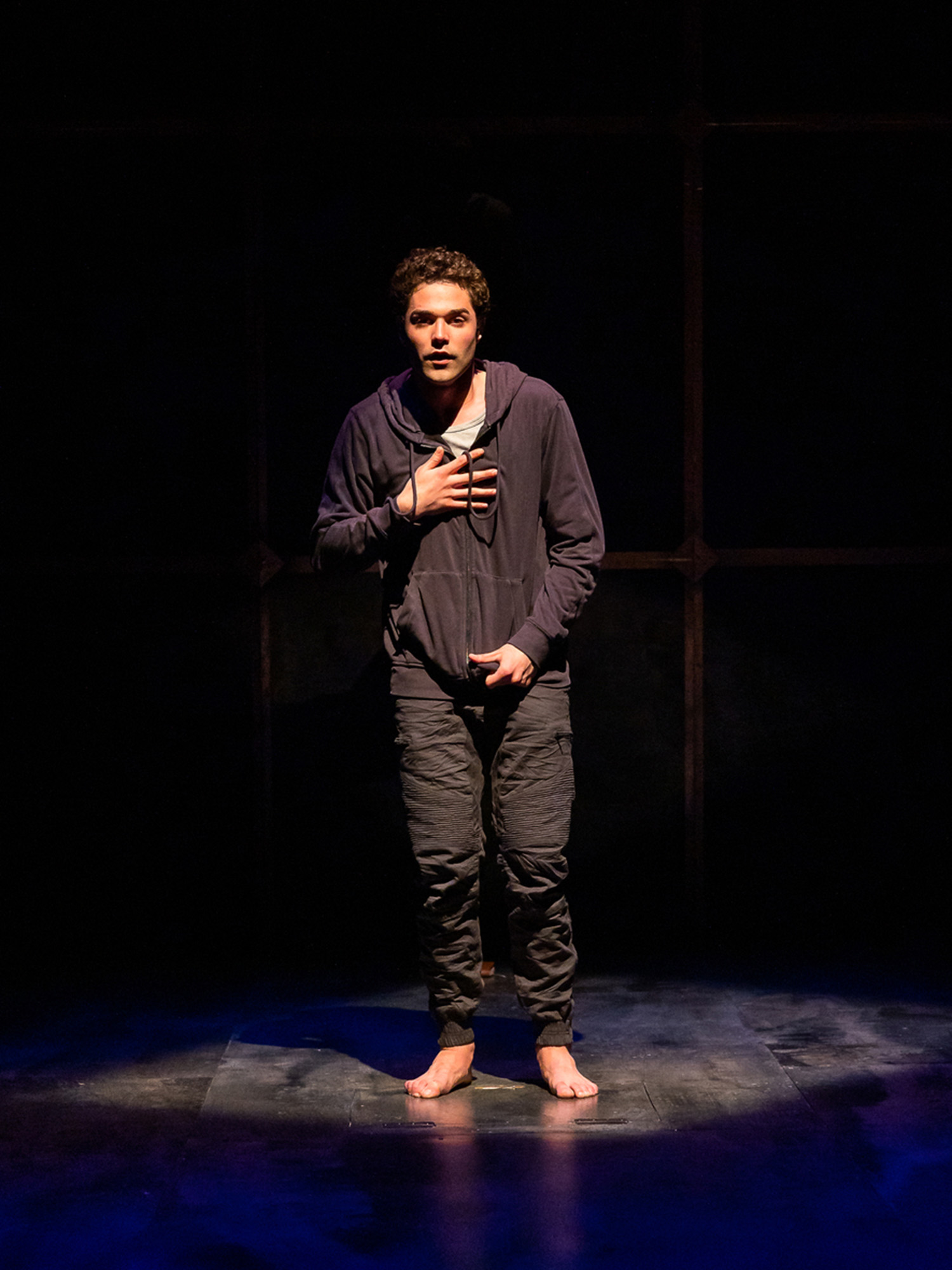 Photo: A young man stands center stage in an isolated stage light. He is barefoot, wearing a black hoodie and black pants. His hand is placed on his chest as he looks out into the audience.