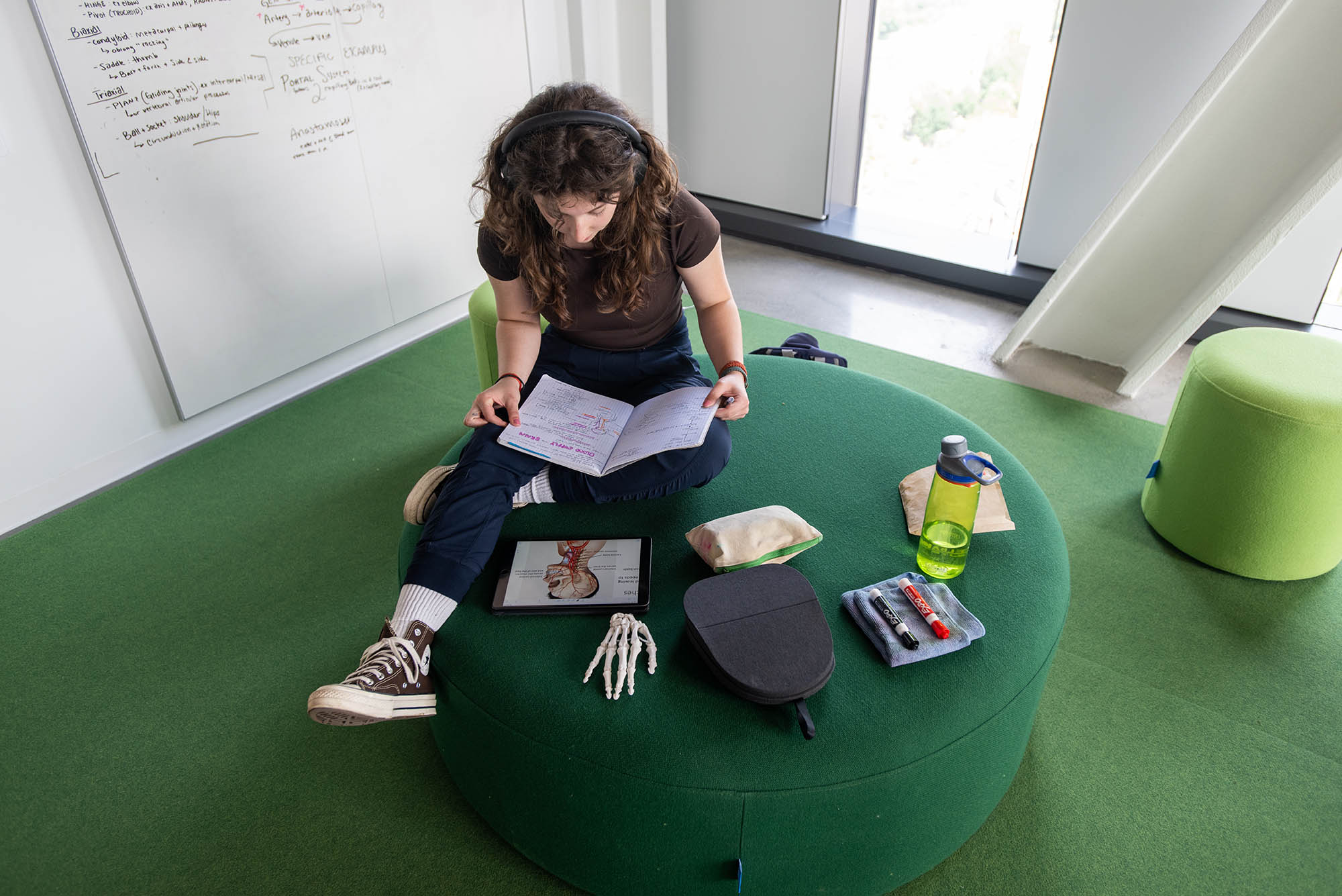 Photo: A young woman sitting on a large green ottoman looks down as she studies. A tablet with an image of the human anatomy is shown and a figure of a skeleton hand is placed in front of her, as well as a water bottle, headphone case, and markers.