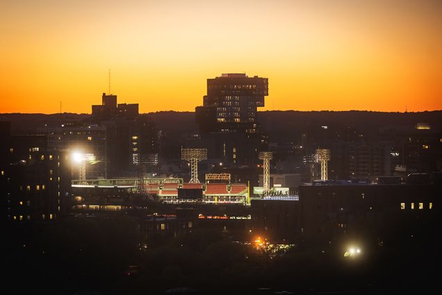 Photo: The sun is setting behind the BU Center of Data Sciences building as a Red Sox game takes place at Fenway park in the foreground. Buildings and scene are shown in shadow as a golden sun set illuminates the scene. The baseball stadium is also lit from within by the stadium lights.