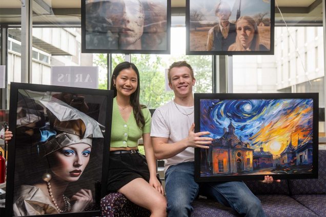 Photo: Two college students, one wearing a black skirt with long black hair and the other wearing a white t shirt with short blonde hair. They are holding up framed pieces of AI art, depicting people and landscapes.