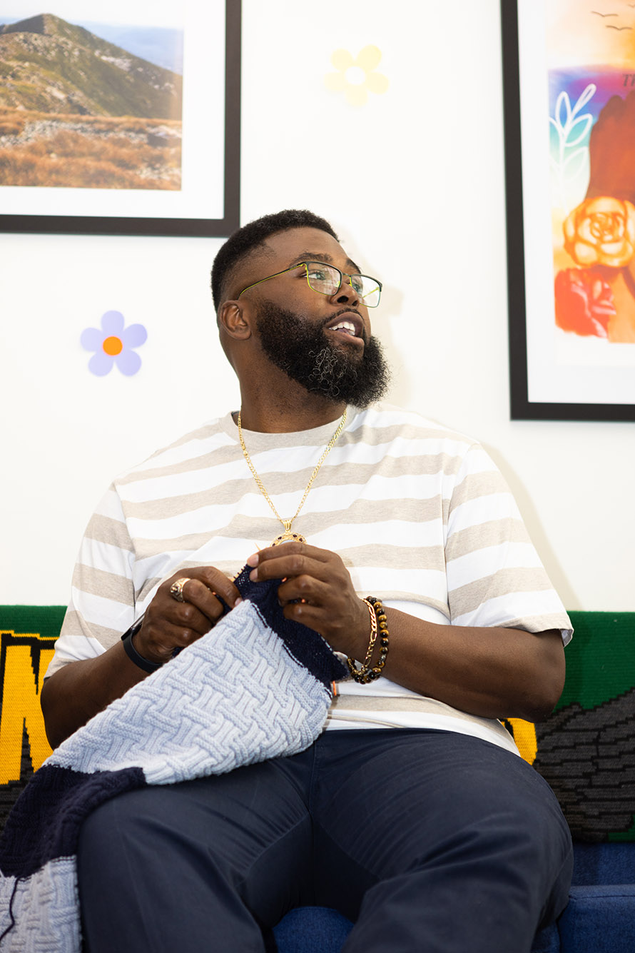 Photo: Anthony Abraham Jack, a Black man wearing glasses, a white and ten striped shirt, and jeans, looks tot he right as he knits with light blue yarn.