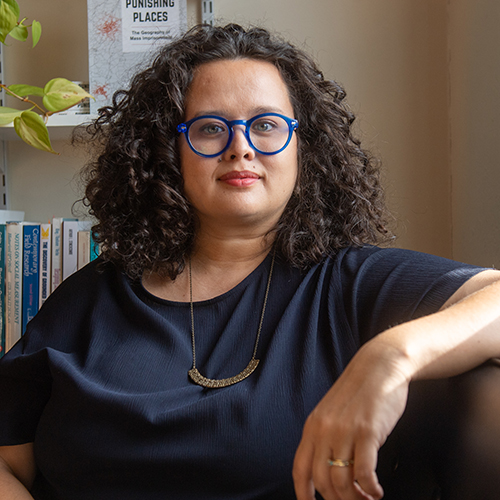 photo of Assistant professor Jessica Simes, a deeply tanned woman with curly dark brown hair and blue oval glasses, sits in her office and faces the camera. Her new book, Punishing Places, is seen on the bookshelf behind her.