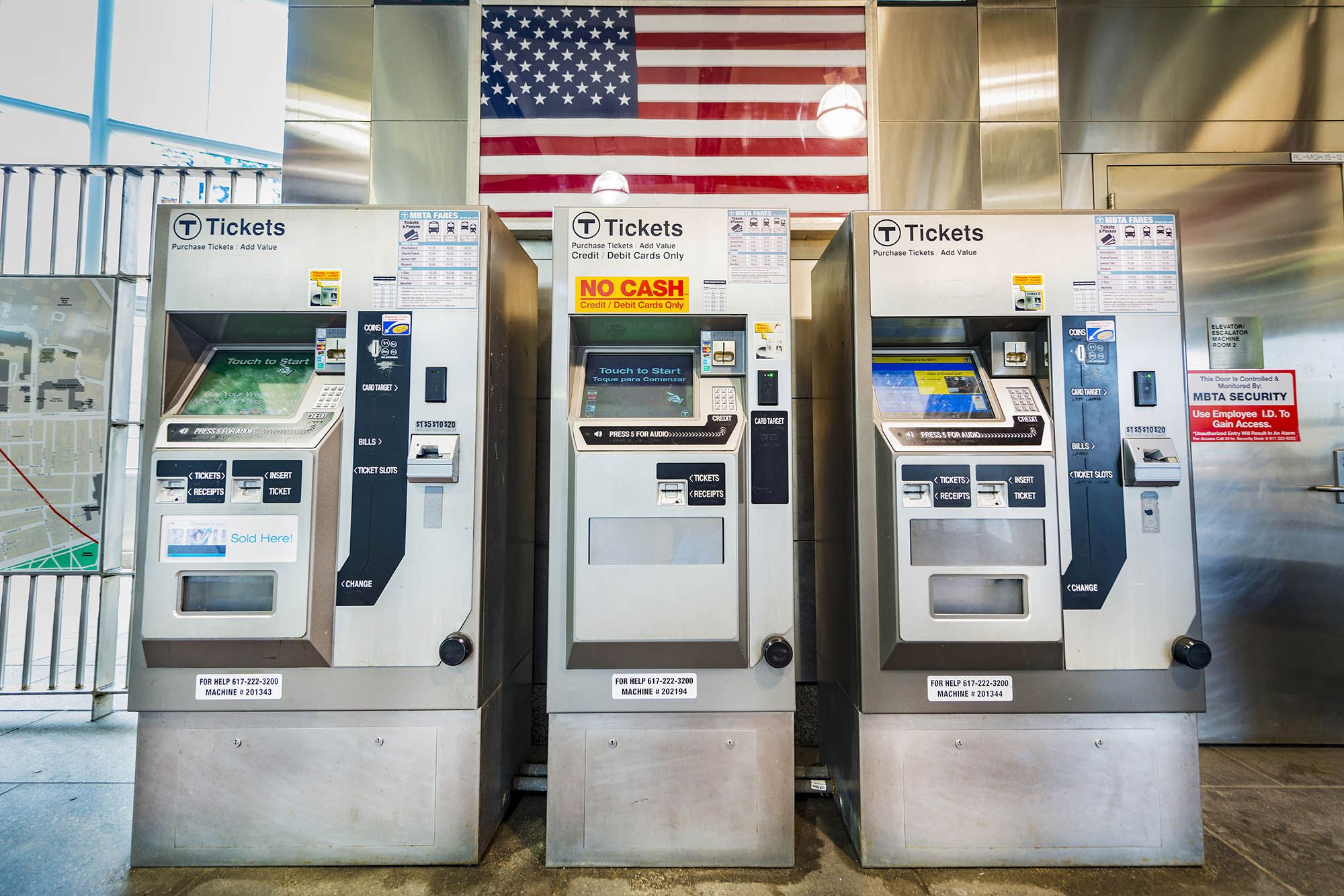 Photo: A row of Ticket Machines Located in Boston Subway. three metal machines for ticketing are shown. The machine in the middle features a yellow sign that reads "no cash".