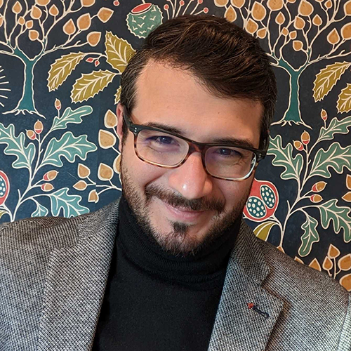 Photo: Renato Mancuso , a white man with curly brown hair and wearing glasses, a black turtleneck sweater, and grey blazer, smiles and poses for a photo in front of a colorful paisley background.