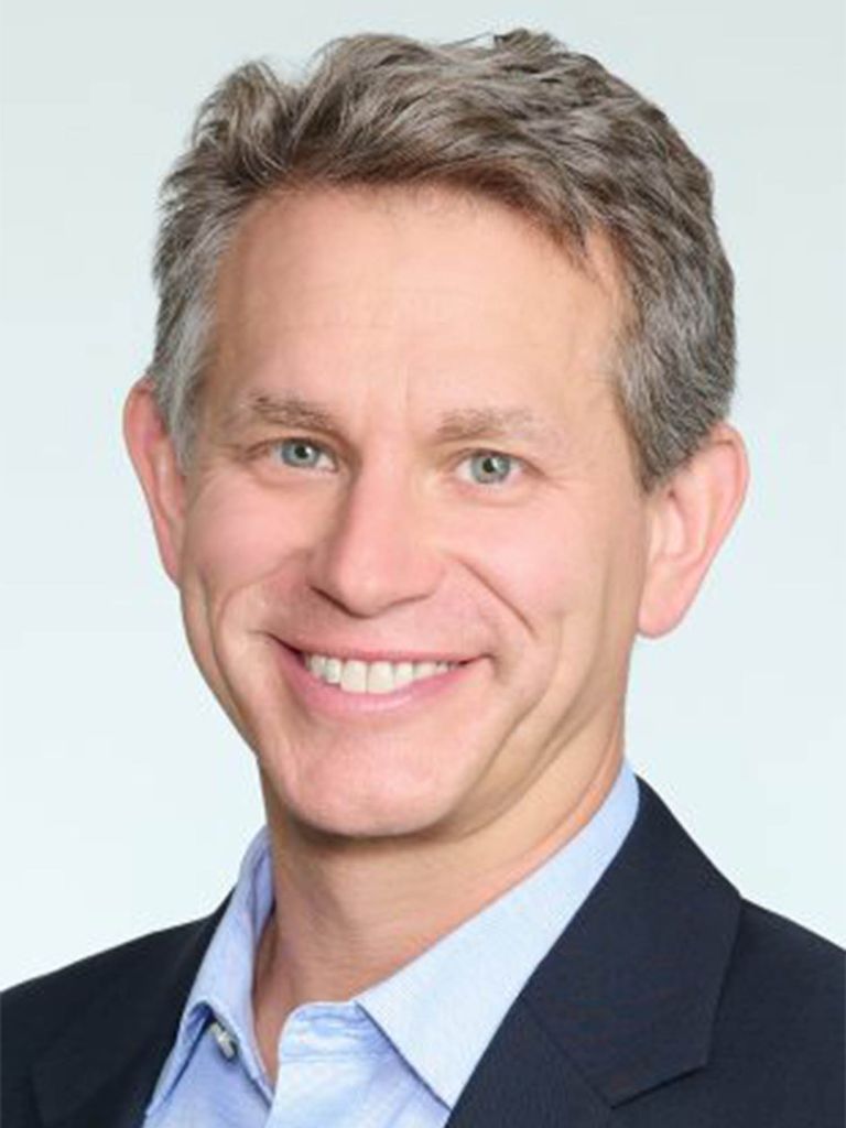 Photo: A headshot of a white man smiling in a black suit with a collared shirt