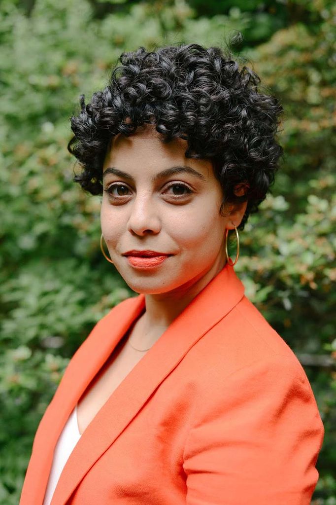 Photo of Heba Gowayed, a light skinned woman who wears gold hoop earrings and lipstick. She has dark, curly hair and wears a pastel orange blazer as she stands outside in front of a bush.