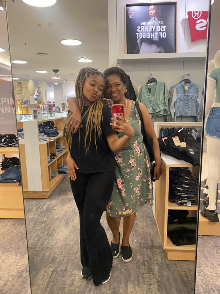 This is a photo by Boston University student Ashley Facey.  She is pictured with her mother as they stand in the mirror taking pictures of themselves.  Ashley is wearing a black dress and has a red phone in her hand.  She purses her lips and has sandy braided hair.  Her mother stands next to her in a floral dress and black shoes.  Her mother's scalp-length curly hair.  She is smiling just behind her daughter. 