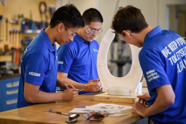 Photo: A group of engineers stand around a hi-tech toilet seat. They are wearing matching blue uniforms and safety glasses.