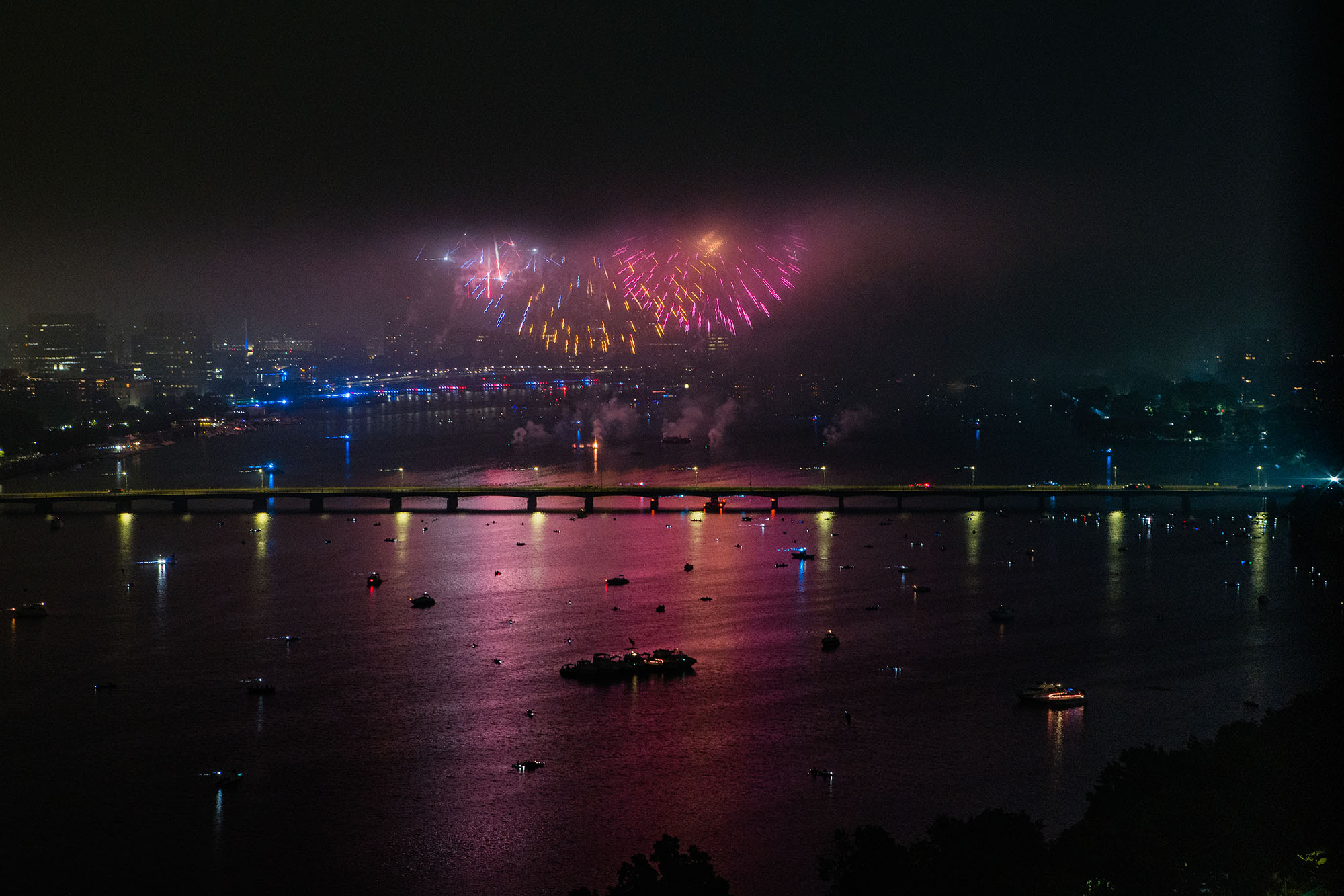 Photo: 4th of July fireworks are shown going off on a dark night above the Charles river. Purple, blue, and yellow fireworks light up the night sky and show the Boston skyline.