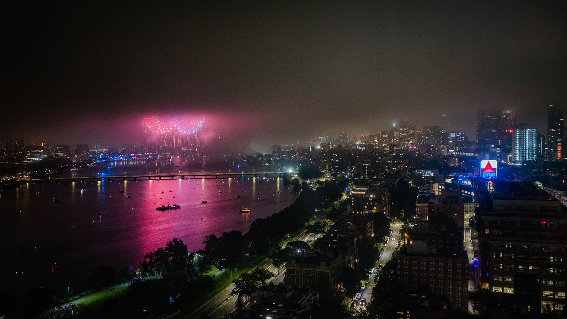 Photo: 4th of July fireworks are shown going off on a dark night above the Charles river. Purple fireworks light up the night sky and show the Boston skyline, BU campus, and bright CITGO sign.