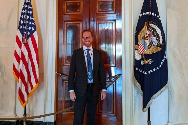 Photo: Goodman, a white man wearing glasses, a white collared shirt, and black suit, stands in front of a large dark wood door. He stands in the center of the US flag and the White house flag.