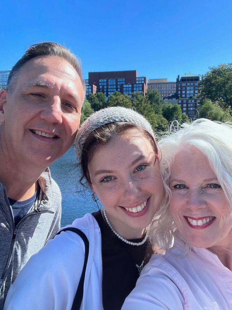 This photo is courtesy of Sophia Harris.  She is with her parents, in front of the water body.  It's a selfie with her in the middle, her mom on the right, and her dad on the left.  The three of them laugh, Sofia wearing a gray headband to push back her brown hair and a black shirt with a white sweatshirt over it.  Her mother wears a pale pink button-up shirt and red lipstick, with white hair.  Her dad has gray hair and wears a gray quarter-zip sweatshirt. 