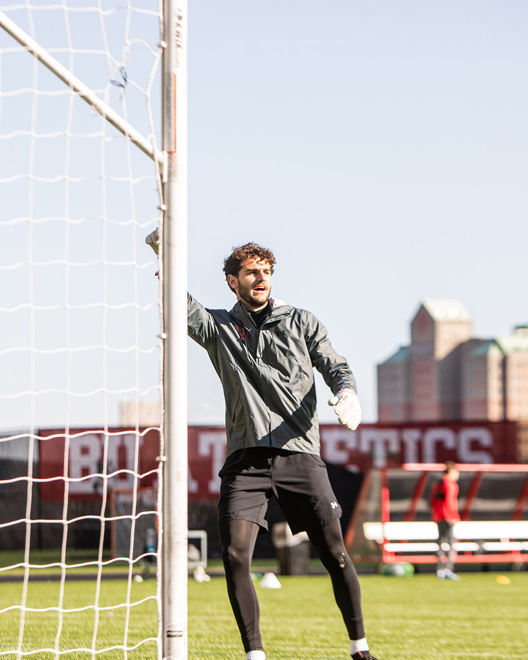 Photo: Action shot of Francesco Montali as he stands and raises his hand in a soccer goal. A young white man with curly brown hair and wearing a grey jacket and black short and spandex, raises his left arm as he looks to the right.