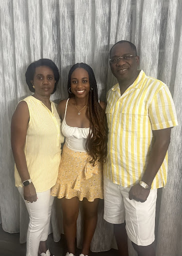 This is a photo of Boston University student Belinda Daniel with her parents. They are all standing in front of a grey curtain wearing yellow and white outfits. Belinda is in the middle and her mother is to the left while her father is on the right. In the middle, she is smiling and wearing a white tank top with a yellow skirt. Her father is also smiling, wearing a yellow striped shirt, glasses, and white shorts. Her mother has a straight face and is wearing a yellow top with white pants.