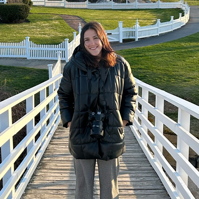 Photo: Alexandra Castro , a young white woman wearing grey sweatpants and black puffer coat, smiles as she poses with hands in coat pockets. A large DSLR hangs from her neck.