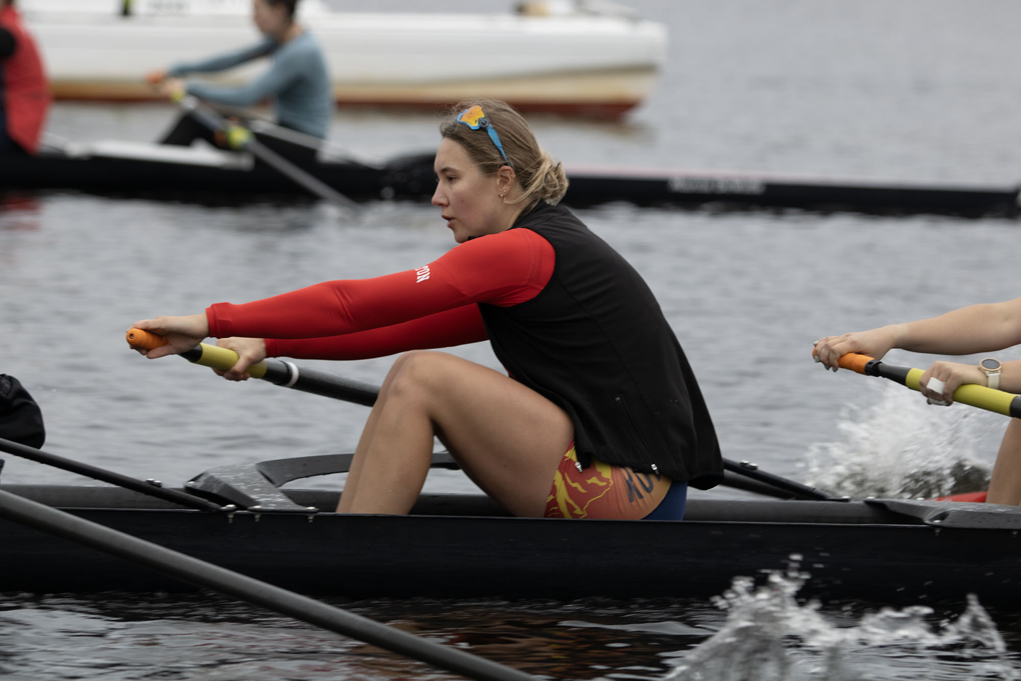 Photo: Action shot of Kat Robbins rowing with her team on the water. A young white woman with hair tied back in a bun and wearing a scarlet red longsleeved, black vest, and swim shorts, is shown mid-row.
