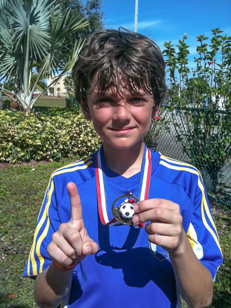 Photo: A young Francesco Montali raises his left index finger in the #1 gesture and shows off a soccer medal in his right hand. A young white boy with curly brown hair and wearing a blue jersey poses for a photo.