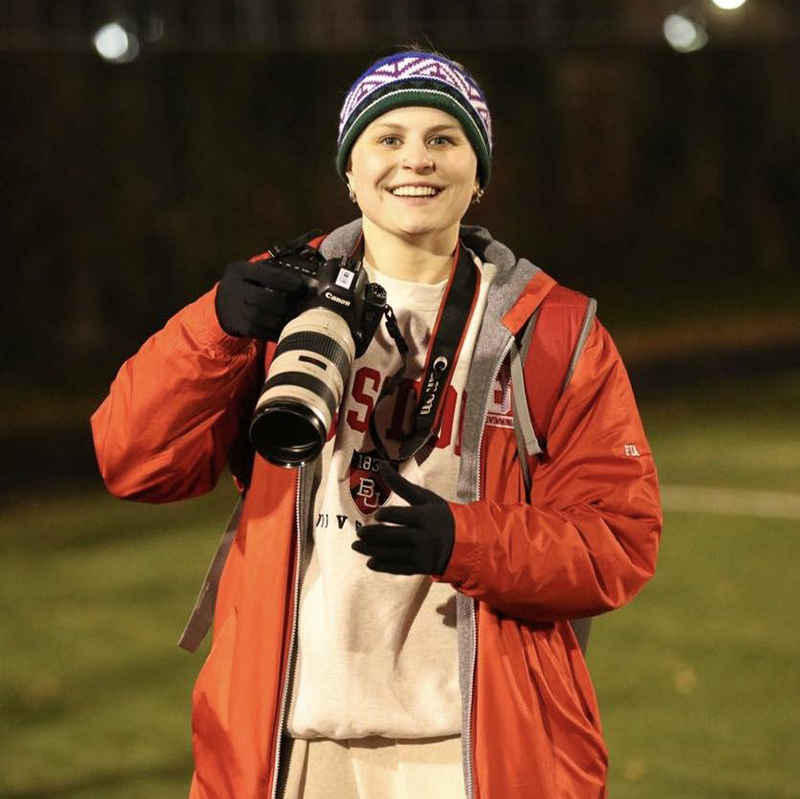 Photo: Sophia McCarty, a young white woman wearing a Boston University grey sweatshirt and pants, red BU coat, wool headband, and black gloves smiles as she holds a DSLR camera with large lens attached to it.