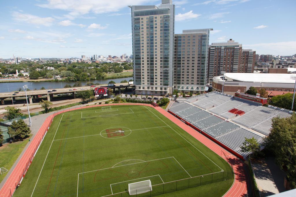 This is a photo of Nickerson Field at Boston University. The field is located adjacent to the Charles River and is a green expanse with a red track looped around it. On the right side, there are sets of bleachers. Next to that, you can see the tall buildings that are BU dorms. 