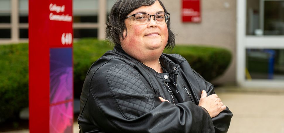 Photo: Joan Donovan, a white woman with short, silver hair and wearing a black top, faux leather jacket, and glasses, poses in front of the BU College of Communication building. Behind her, a red sign reads "College of Communication".