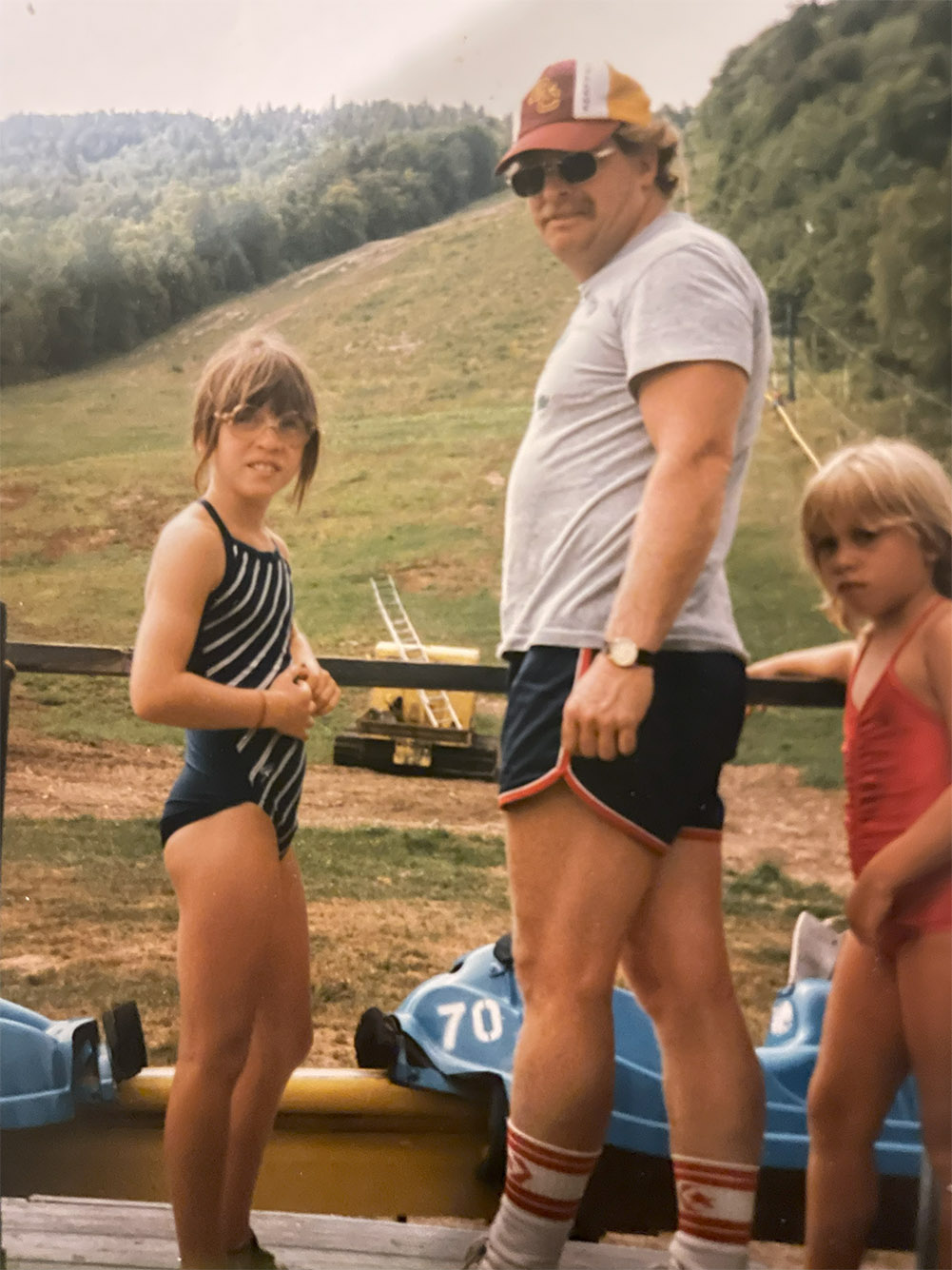 Photo: Older photo of a young Laurie Craigen (left) next to her father. A young child in a black and white full swimsuit stands and looks over her shoulder at the camera. To her right an older man wearing sunglasses, a hat, grey shirt, and black shirts, looks over his shoulder to the camera as well.