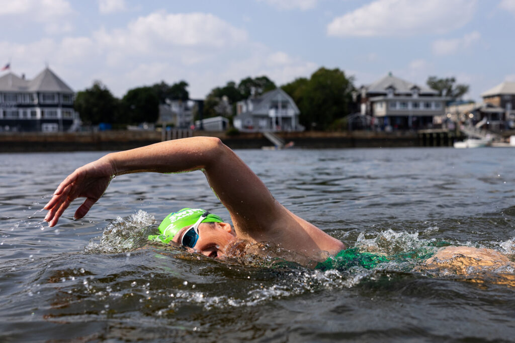 Photo: Action shot of Laurie Craigen, a white woman wearing a full green and black patterned swimsuit, green swim cap, and goggles. She is shown swimming in the Boston Harbor.