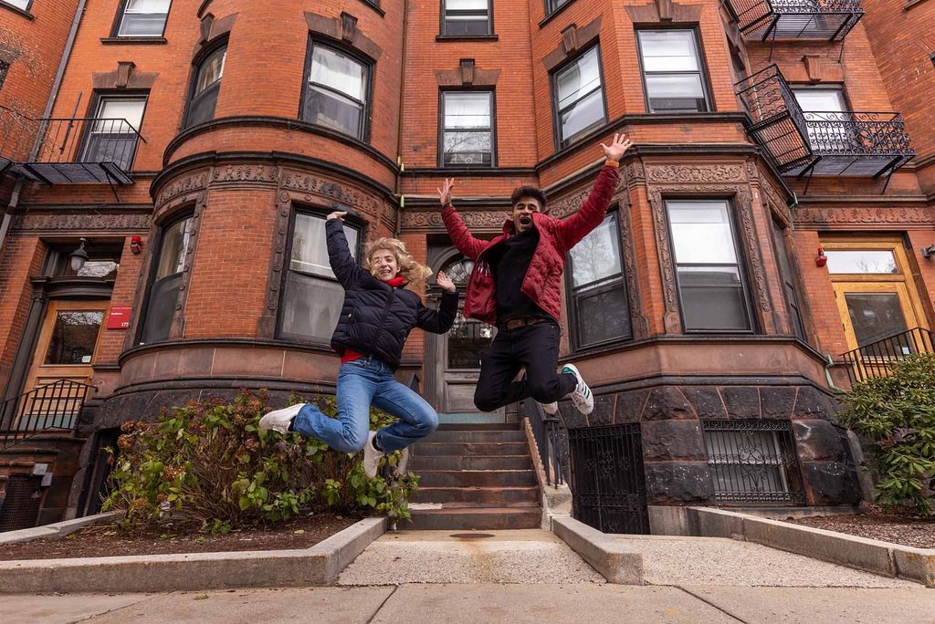 This is a photo in front of the Brown Stone dorms at Boston University on east campus. There are two individuals on the side walk in front of the brownstones and they are jumping in the air smiling and flailing their arms out. A blonde girl in a black jacket and blue jeans is on the left while a brunette man in a red jacket, black shirt, and black jeans is on the left. 