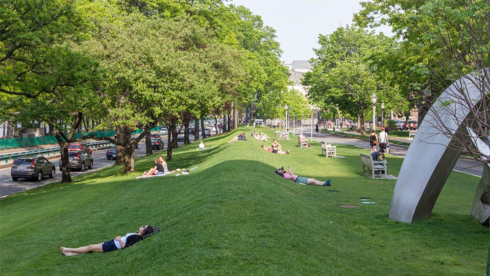 This is a photo of what Boston University students refer to as the 'BU Beach.' It is not an actual sandy beach but it is a place where students lay down and hear the noises of the highway, which is supposed to sound like ocean waves. The photo shows green grass with green trees lining the outside of parameter. To the left and the right, there is road that has cars lined on the sides of it. Across the green grass, there are students scattered about, laying down. Most of them are reading or looking up at the sky. 