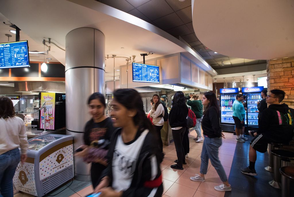 This is a blurry action photo taken at a Boston University dining hall. There are multiple students that are blurry and walking all different ways. The person closest to the camera is wearing a white and black shirt and is walking to the left of the shot. In the back, there is a blurry mess of individuals, heading towards the food menus and seats. The food menus are displayed on two different TVs. There is also a vending machine and a reach in freezer.