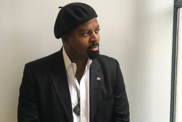 Photo: A black man in a beret and a collared shit with a suit jacket poses for a formal photo
