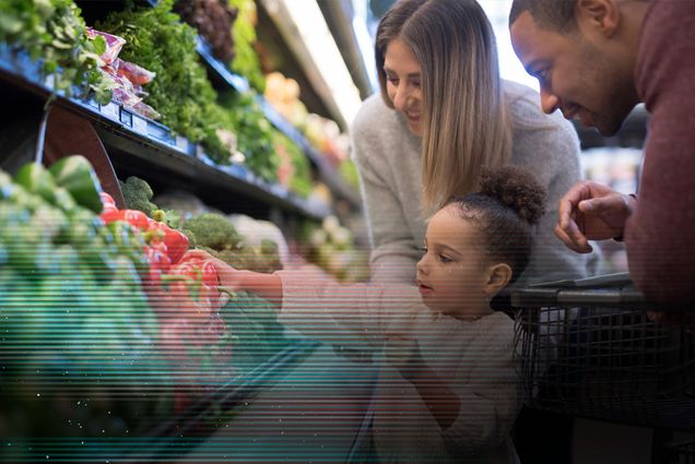 Photo: A pre-school age girl helps her parents pick out veggies in the produce section at the grocery store. She is reaching for a red bell pepper. Her mother is Caucasian and her father is African-American.