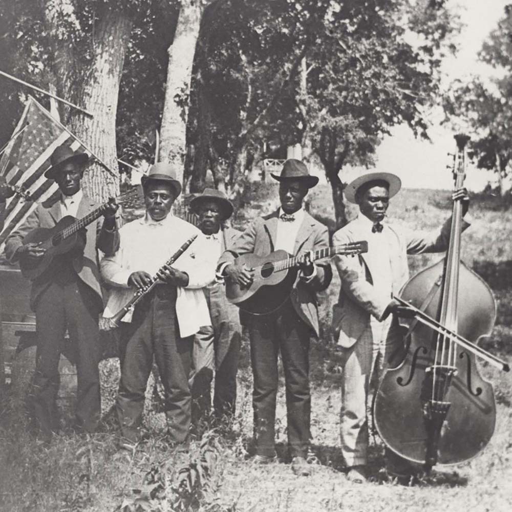 Black and white photograph of a band of African-American playing at Emancipation Day celebration in June 19, 1900, held in “East Woods” on East 24th Street in Austin. There are 6 men in the band, they all wear suits and hats and hold their instruments, including a fiddle, guitar, clarinet, and upright bass. There are trees and an American flag hanging behind them.