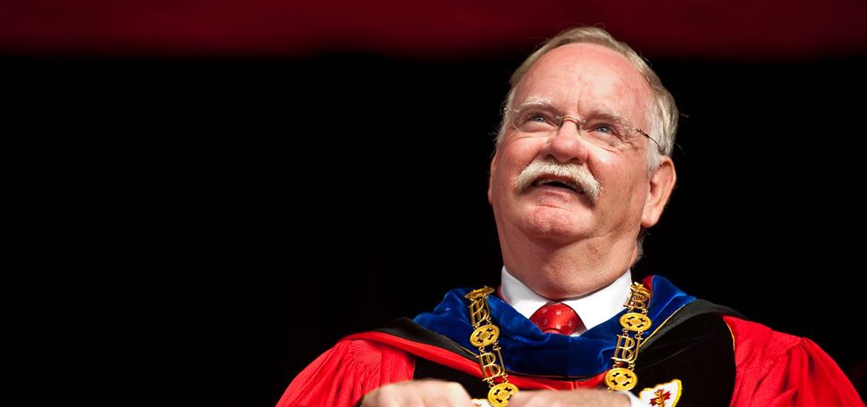 Photo of President Robert Brown in red ceremonial regalia. He has a blue color and wears a gold BU necklace. He is seated, and balances a red folder in his lap and smiles as he looks up into the middle distance. Brown is an older White man with glasses and short gray hair and a mustache.