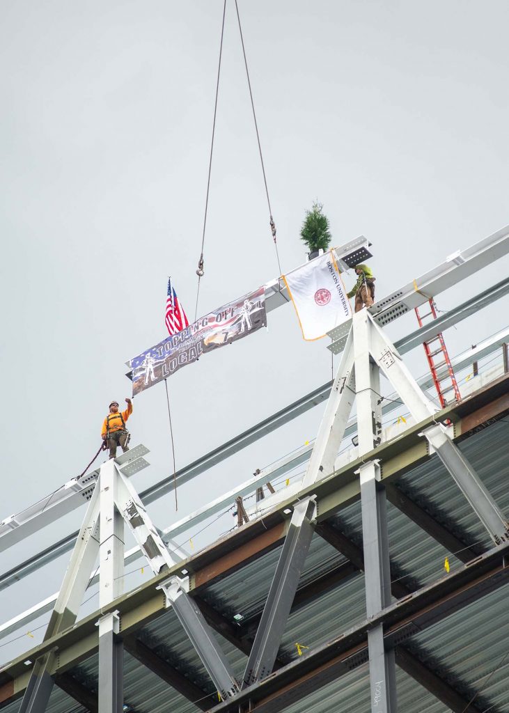 .Covered in signatures, the final beam is put into place during the Topping Off ceremony. A large beam is guided into place by two construction workers on top of the building. The sky is grayish blue.
