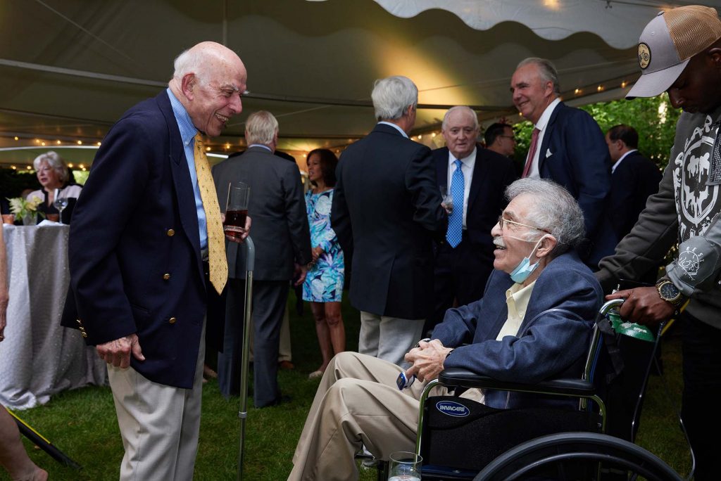 Aram, at right, is a light-skinned Armenian man with gray hair. He wears a blue suit and yellow tie, holds a glass and a cane in his left hand and smiles as he talks to his life long friend. Edward, at left, is a light-skinned Armenian man with gray hair. He smiles and sits in a wheelchair, wears a blue suit jacket, and wears a face mask underneath his chin. A young man of color holds his wheelchair. Others dressed in formal attire can be seen talking underneath a white tent in the background.