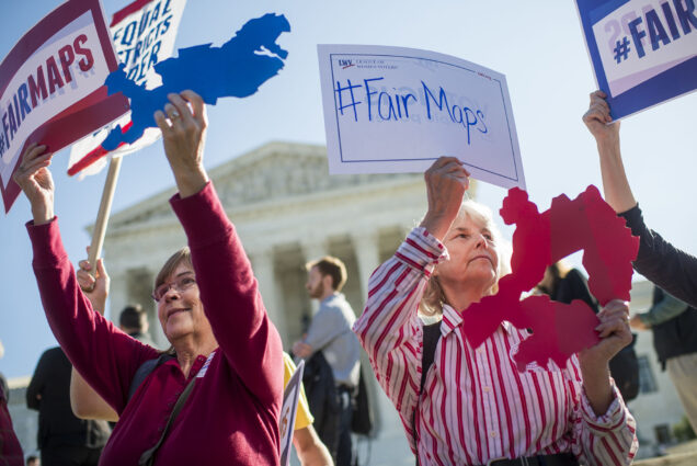 Photo: Protestors wearing red and white clothes hold up signs that read "#FairMaps" as they stand in front of the US Supreme Court building.