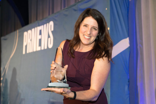 Photo: Amy Shanler, a white woman with long dark brown hair and wearing a burgundy dress, smiles and poses with a glass trophy in front of a blue backdrop that reads "PRNEWS".