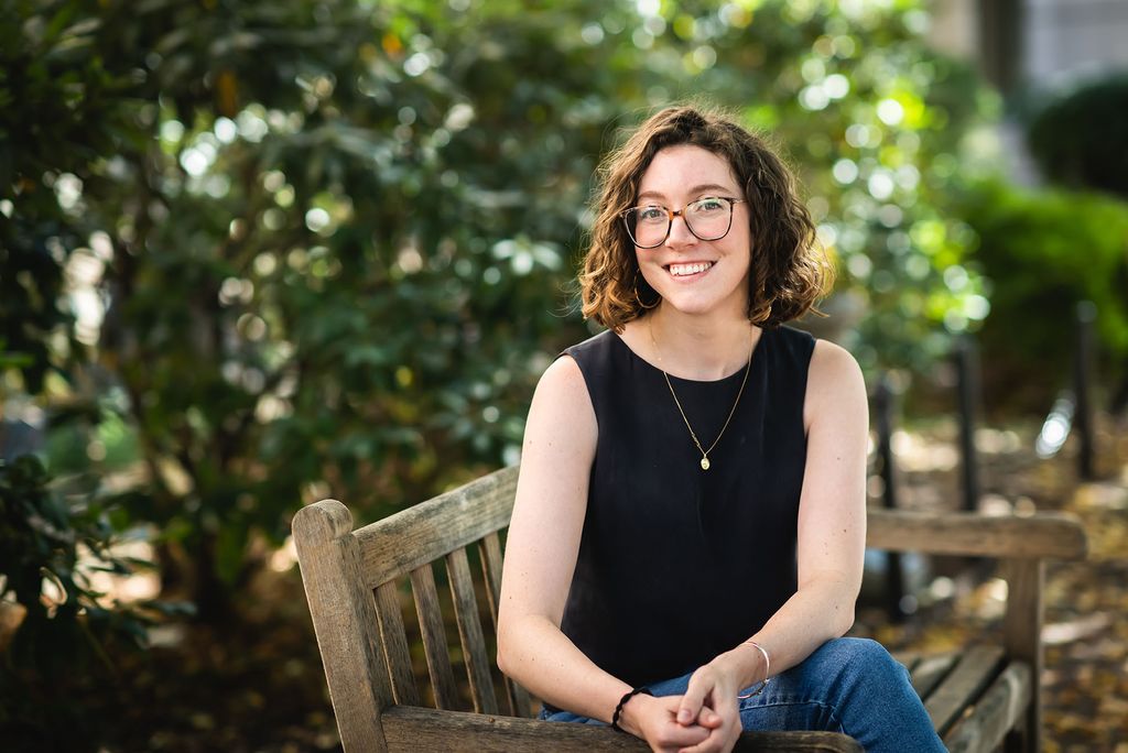 Photo: Annie Goodridge, a young white woman with short, brown, curly hair and wearing glasses, a black blouse, and jeans, smiles and poses on a wooden bench in a lush, green park area.